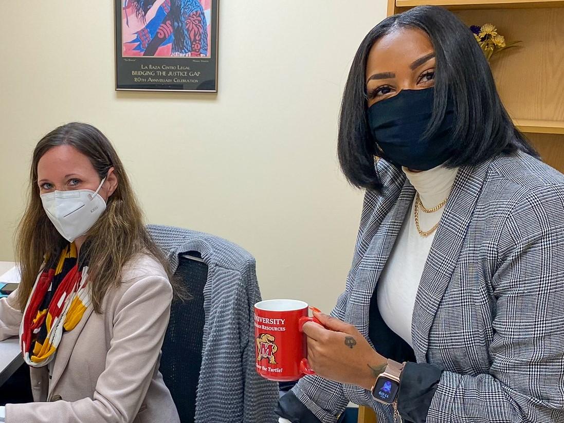 Two female UHR staff wearing masks and facing forward. One holds a mug of coffee.